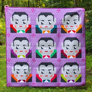 May include: A handmade quilt featuring a repeating pattern of pixelated vampire faces on a lavender background. Each vampire has a different colored bow tie. The quilt is displayed outdoors in front of a green forest.
