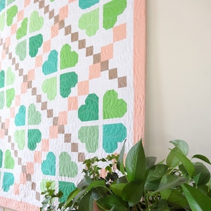 May include: A white quilt with a pink border and a pattern of green four-leaf clovers. The quilt is hanging on a wall next to a potted plant with green leaves.
