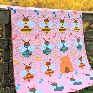 Peut inclure: Une courtepointe rose avec une ruche et de nombreuses abeilles de différentes couleurs, notamment le brun, le jaune, le bleu et l'orange. La courtepointe est accrochée à une clôture en bois avec un arrière-plan flou de fleurs jaunes.