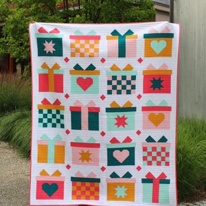 May include: A white quilt with a repeating pattern of colorful gift boxes. Each box has a different design, including stars, hearts, and checkered patterns.