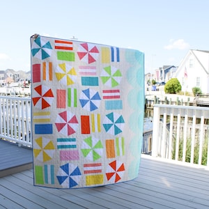May include: A colorful quilt with a beach theme. The quilt is made up of squares with different patterns, including stripes, polka dots, and umbrellas. The quilt is hanging on a railing overlooking a body of water.
