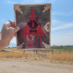 Puede incluir: Un libro rojo, negro y dorado con un s&iacute;mbolo triangular y el texto "The Book of Bill" en la portada.