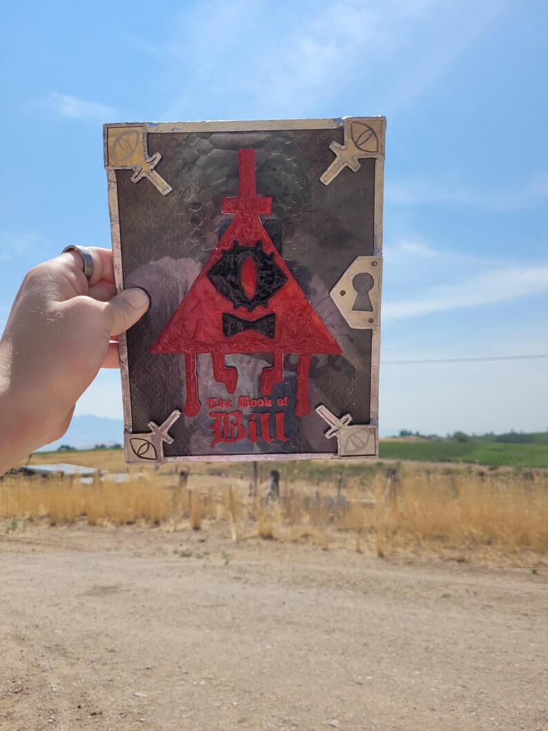 Puede incluir: Un libro rojo, negro y dorado con un s&iacute;mbolo triangular y el texto "The Book of Bill" en la portada.