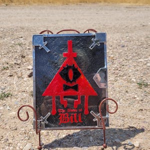 Puede incluir: Una representaci&oacute;n en vitral de Bill Cipher del programa Gravity Falls. La imagen est&aacute; enmarcada en un marco de metal negro y colocada en un soporte de metal. La imagen presenta un tri&aacute;ngulo rojo con un solo ojo amarillo, una corbata de mo&ntilde;o negra y el texto "The Book of Bill" en rojo.