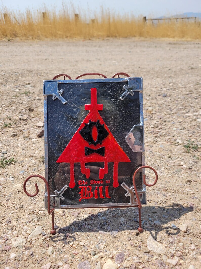 Puede incluir: Una representaci&oacute;n en vitral de Bill Cipher del programa Gravity Falls. La imagen est&aacute; enmarcada en un marco de metal negro y colocada en un soporte de metal. La imagen presenta un tri&aacute;ngulo rojo con un solo ojo amarillo, una corbata de mo&ntilde;o negra y el texto "The Book of Bill" en rojo.