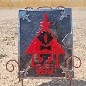 Puede incluir: Una representaci&oacute;n en vitral de Bill Cipher, un personaje de la serie animada Gravity Falls. La imagen est&aacute; sobre un fondo negro y presenta un tri&aacute;ngulo rojo con un ojo amarillo y una pajarita negra. El texto "The Book of Bill" est&aacute; escrito en letras rojas y negras debajo del tri&aacute;ngulo.