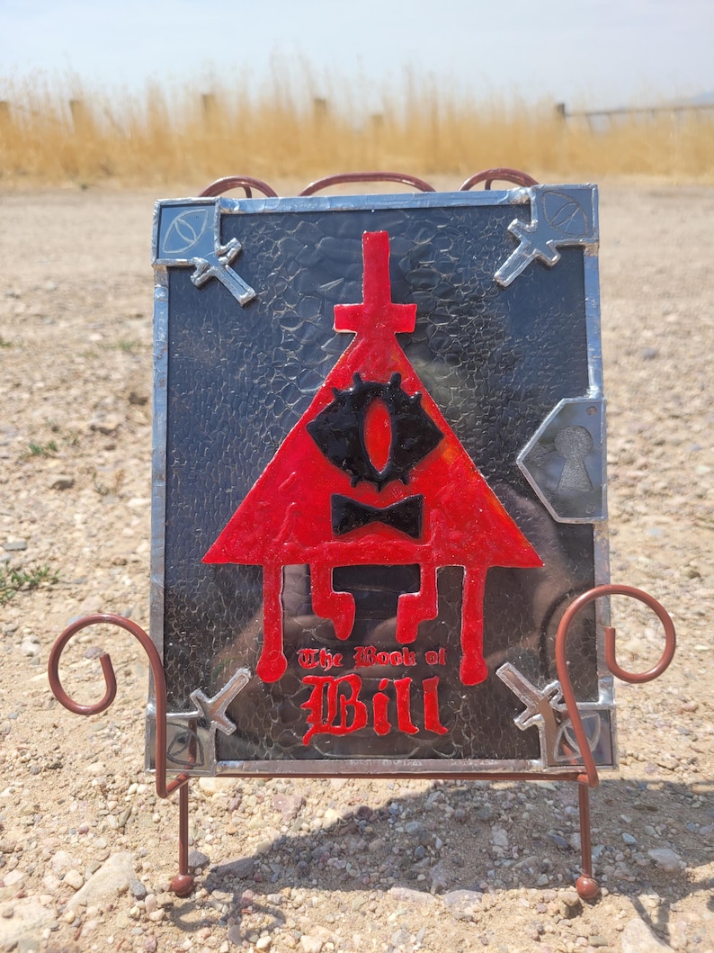 Puede incluir: Una representaci&oacute;n en vitral de Bill Cipher, un personaje de la serie animada Gravity Falls. La imagen est&aacute; sobre un fondo negro y presenta un tri&aacute;ngulo rojo con un ojo amarillo y una pajarita negra. El texto "The Book of Bill" est&aacute; escrito en letras rojas y negras debajo del tri&aacute;ngulo.