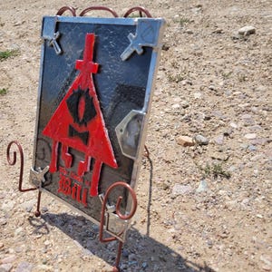 Puede incluir: Una ventana de vitral con un tri&aacute;ngulo rojo con un ojo negro y una cruz en la parte superior. El tri&aacute;ngulo est&aacute; rodeado de un fondo negro con detalles plateados. El texto "The Author of All Evil" y "Bill" se puede ver en la parte inferior de la ventana.