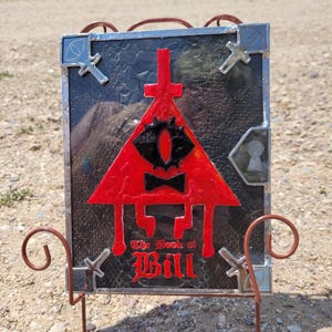 Puede incluir: Una representaci&oacute;n en vitral de Bill Cipher, un demonio triangular de la serie animada Gravity Falls. La imagen presenta un tri&aacute;ngulo rojo con un solo ojo, una pajarita y el texto "The Book of Bill".