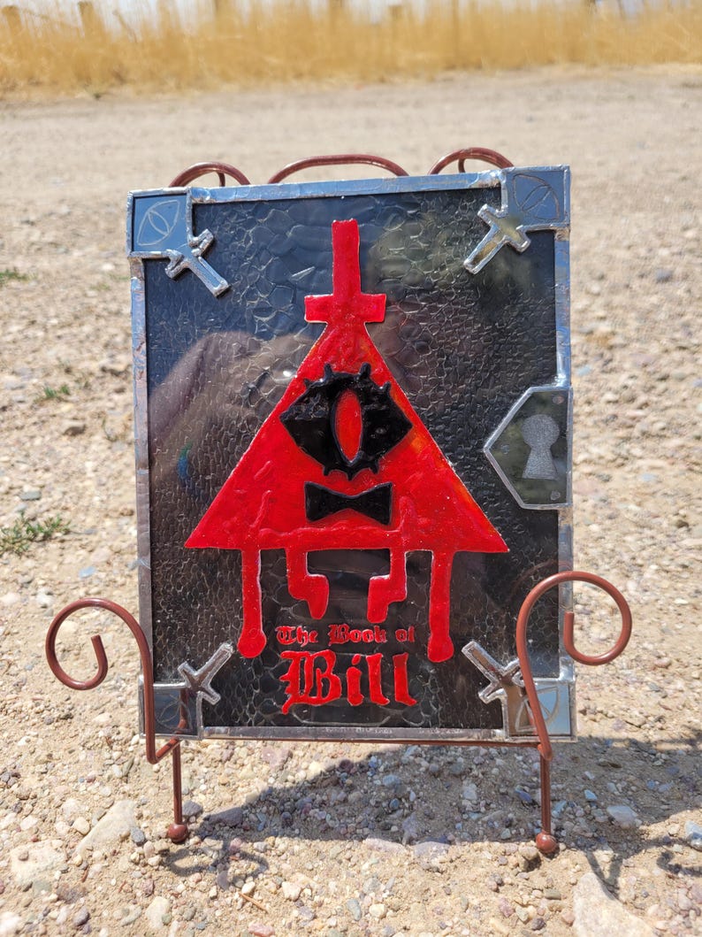 Puede incluir: Una representaci&oacute;n en vitral de Bill Cipher, un demonio triangular de la serie animada Gravity Falls. La imagen presenta un tri&aacute;ngulo rojo con un solo ojo, una pajarita y el texto "The Book of Bill".