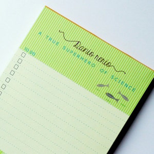 Peut inclure: Un bloc-notes rayé vert et blanc avec le texte "Danio rerio SUPERHERO OF SCIENCE" et une liste de choses à faire avec des cases à cocher. Le bloc-notes a une bordure verte et une petite illustration de poisson.