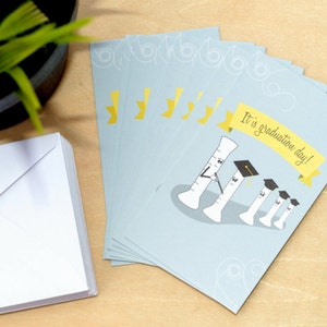 May include: A stack of blue graduation cards with a yellow banner that reads "It is graduation day!" and a cartoon illustration of four graduated test tubes.
