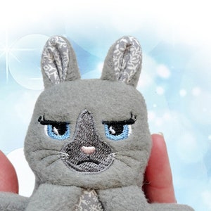 May include: Gray plush toy rabbit with embroidered blue eyes and a floral pattern on its chest. The rabbit is sitting in a hand.
