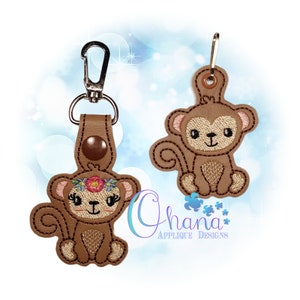 以下が含まれることがあります： 2つの茶色の猿のキーホルダー。1つは花の冠をつけた猿、もう1つはシンプルなデザインの猿です。どちらも茶色の革の裏地と金属製の金具が付いています。「Ohana Applique Designs」の文字が見えます。