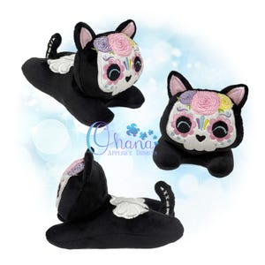 May include: A black plush cat toy featuring a white face mask with embroidered pink, purple, and yellow floral details around the ears and eyes. The cat has a white wing-like design on its back and a small tail. The text Ohana Applique Designs is visible.