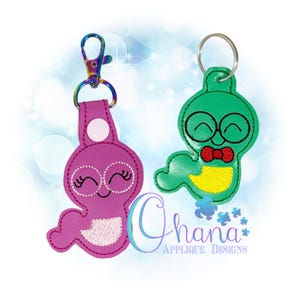 Puede incluir: Dos llaveros con diseños de gusanos de dibujos animados. Uno es morado con un cierre plateado, y el otro es verde con un llavero plateado. Ambos gusanos tienen gafas y caras sonrientes. El gusano verde tiene una pajarita roja y una barriga amarilla.