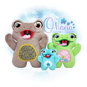 Könnte beinhalten: Drei Plüschfrosch-Spielzeuge in verschiedenen Größen. Der größte ist hellbraun mit einem gelb-schwarzen Bauch, der mittlere ist grün mit einem rosa Schmetterlingsdesign und der kleinste ist blau mit einem weißen Stern. Jeder hat ein lächelndes Gesicht und eine herausgestreckte Zunge.