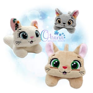 May include: Three plush cat toys featuring embroidered details, large expressive eyes, and small pink noses. One is cream-colored with floral-patterned ears, one is light gray, and one is tan. The text Ohana Applique Designs is visible.