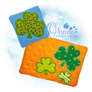 Pode incluir: Dois quadrados de tecido, um laranja e outro azul, com desenhos de trevo bordados. Os trevos são verdes com padrões diferentes. O quadrado laranja tem três trevos e o quadrado azul tem um trevo. O texto "Ghana Applique Designs" é visível no fundo.