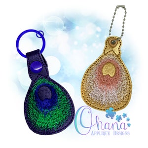 Puede incluir: Dos llaveros con diseños bordados. Un llavero es morado con un diseño de pluma de pavo real verde y azul. El otro llavero es dorado con un diseño de pluma rosa y blanco. Ambos llaveros tienen una cadena plateada.