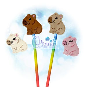 Könnte beinhalten: Vier Stifteaufsätze in Form von Wasserschweinen in Braun-, Beige-, Creme- und Rosatönen. Jeder Aufsatz ist an einem Stift mit einem Regenbogenverlauf befestigt. Das Bild enthält den Text "Ohana Applique Designs".