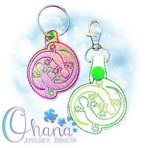 May include: Two round keychains with a green gecko design. One keychain is pink with a holographic finish and the other is white with a green gecko design.
