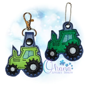 May include: Green and black tractor keyring with a blue leather strap and a silver metal clasp. The tractor has a green body, black wheels, and a white window.