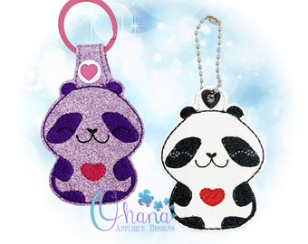 May include: Two panda keychains, one purple with glitter and one black and white. Both pandas are holding a red heart. The purple panda keychain has a pink heart on top and a pink ring. The black and white panda keychain has a black heart on top and a silver chain.