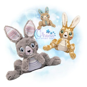 Pode incluir: Três brinquedos de pelúcia de coelho com detalhes bordados. Os coelhos são cinza, marrom e bege com tecido listrado colorido em suas orelhas e corpos. Os coelhos estão sentados em um fundo azul e branco.