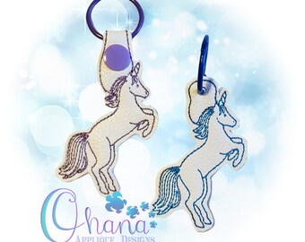May include: Two embroidered unicorn keychains. One keychain is white with a purple button and a black key ring. The other keychain is white with a blue tail and a black key ring.