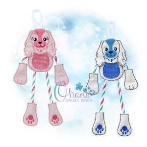 May include: Two embroidered dog ornaments with candy cane legs. The dog on the left is pink and white, and the dog on the right is blue and white.