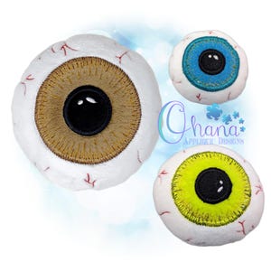 May include: Three plush, embroidered eye designs. One eye has a brown iris, another has a blue iris, and the third has a yellow iris. Each eye has a black pupil and red veins. The text "Ohana Applique Designs" is also visible.