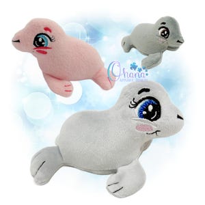 Puede incluir: Tres juguetes de peluche con forma de foca que presentan detalles bordados, incluyendo grandes ojos azules con pestañas largas y mejillas rosadas. Las focas son de tela rosa y gris claro. El logo Ohana Applique Designs es visible.