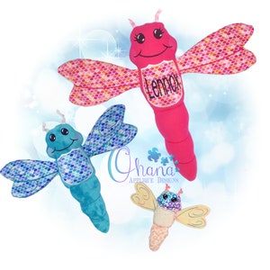 Puede incluir: Tres juguetes de peluche con forma de libélula en tonos rosa, azul y crema. La libélula rosa tiene el nombre "Lennox" en su cuerpo. Las alas presentan un patrón de escamas. Los juguetes tienen caras sonrientes y están diseñados para niños.