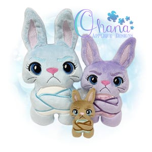May include: Three plush bunny toys in light blue, purple, and brown. Each bunny has a grumpy expression, large blue eyes, and pink accents. The bunnies are wrapped in fabric and have long ears. The image includes the text "Ohana Applique Designs".
