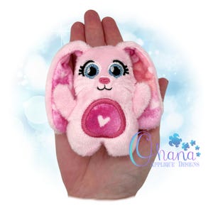 Op de afbeelding: Een roze pluche konijn met een hart op zijn borst. Het konijn heeft geborduurde ogen en een roze neus. Het konijn wordt in een hand gehouden.