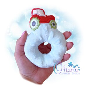May include: A soft, light blue donut-shaped toy with a red and white tractor applique. The tractor features green and grey details. The toy is plush and fluffy, suitable for babies or young children.
