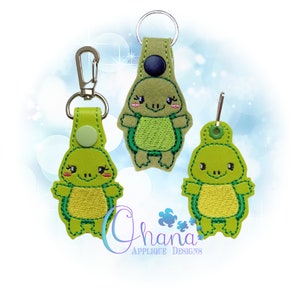 以下が含まれることがあります： 刺繍のディテールが施された緑色のカメのキーホルダーが3つ。各キーホルダーには、銀色の金属リングと緑色の革のストラップが付いています。カメは黒い目とピンクの頬をしています。