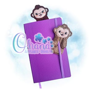 May include: A purple notebook with a matching elastic band and a ribbon bookmark. Two monkey-shaped felt bookmarks are attached to the notebook. The text "Ohana Applique Designs" is visible on the notebook.