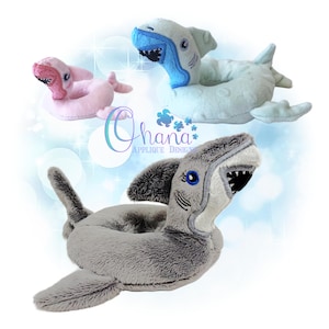 May include: Three plush shark-shaped toys in shades of pink, blue, and gray. The sharks have open mouths with visible teeth and embroidered details. The image includes the text "Ohana Applique Design".