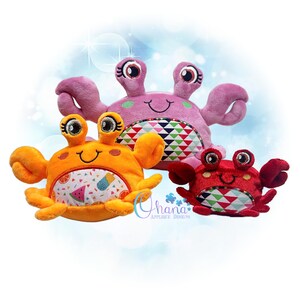 Pode incluir: Três brinquedos de pelúcia em forma de caranguejo nas cores laranja, rosa e vermelho. Cada caranguejo tem olhos grandes, rostos sorridentes e barrigas estampadas. O caranguejo laranja tem uma barriga com tema de frutas. O caranguejo rosa está ao fundo.