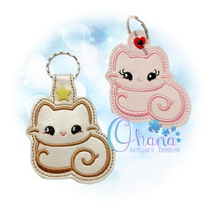 Peut inclure: Deux porte-clés en forme de chat avec des anneaux métalliques. L'un est crème avec une broderie marron et une étoile jaune. L'autre est rose clair avec une broderie rose et un petit cœur rouge. Le texte Ohana Applique Designs est visible.