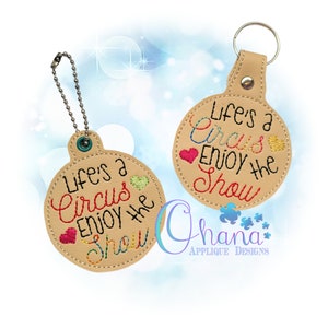 May include: Two brown leather keychains with a circle-shaped tag. The tag has colorful embroidery that reads "Life's a Circus Enjoy the Show".