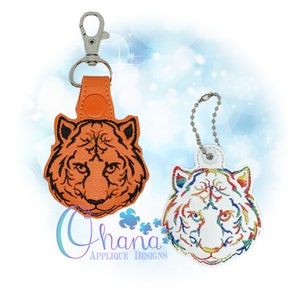 May include: Two embroidered tiger head keychains. One is orange with black details and the other is white with rainbow details. Both have a silver chain.