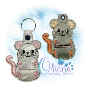 May include: Two mouse-shaped keychains. One is silver with pink accents and a coin pocket. The other is gray with orange accents. Both have embroidered details, including whiskers and paws. The keychains are designed with a cute, cartoonish style.