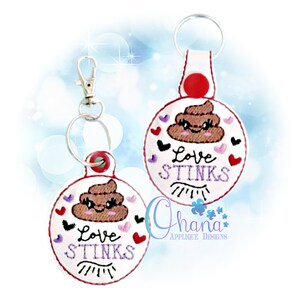 May include: Two round keychains with a brown poop emoji with a smiling face and eyelashes. The keychains are white with red hearts and the text "Love Stinks" in purple.