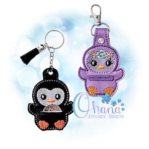 Puede incluir: Dos llaveros con forma de pingüino. Uno es negro con pies naranjas y una moneda de plata en su bolsillo. El otro es morado con una corona floral y pies naranjas. Ambos tienen llaveros y cierres metálicos.