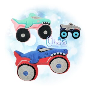 May include: Three plush monster truck toys with shark designs. The trucks are blue, green, and red with black, white, and gray details. The trucks have large wheels and are in a white background.