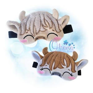 Op de afbeelding: Twee slaapmaskers in de vorm van een Schotse Hooglandkoe. Eén masker is lichtbruin en de andere bruin. Beide maskers hebben zwarte elastische banden, roze wangen en lachende gezichten. De maskers zijn gemaakt van zachte stof.