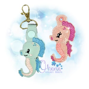 May include: Two embroidered seahorse keychains, one in light blue and pink, the other in pink and peach. Both have a cute, cartoon-like face and a gold key ring.